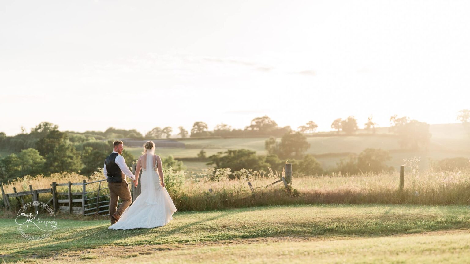 A bride and groom holding hands and walking in a grassy field during sunset, with a rural landscape in the background.
