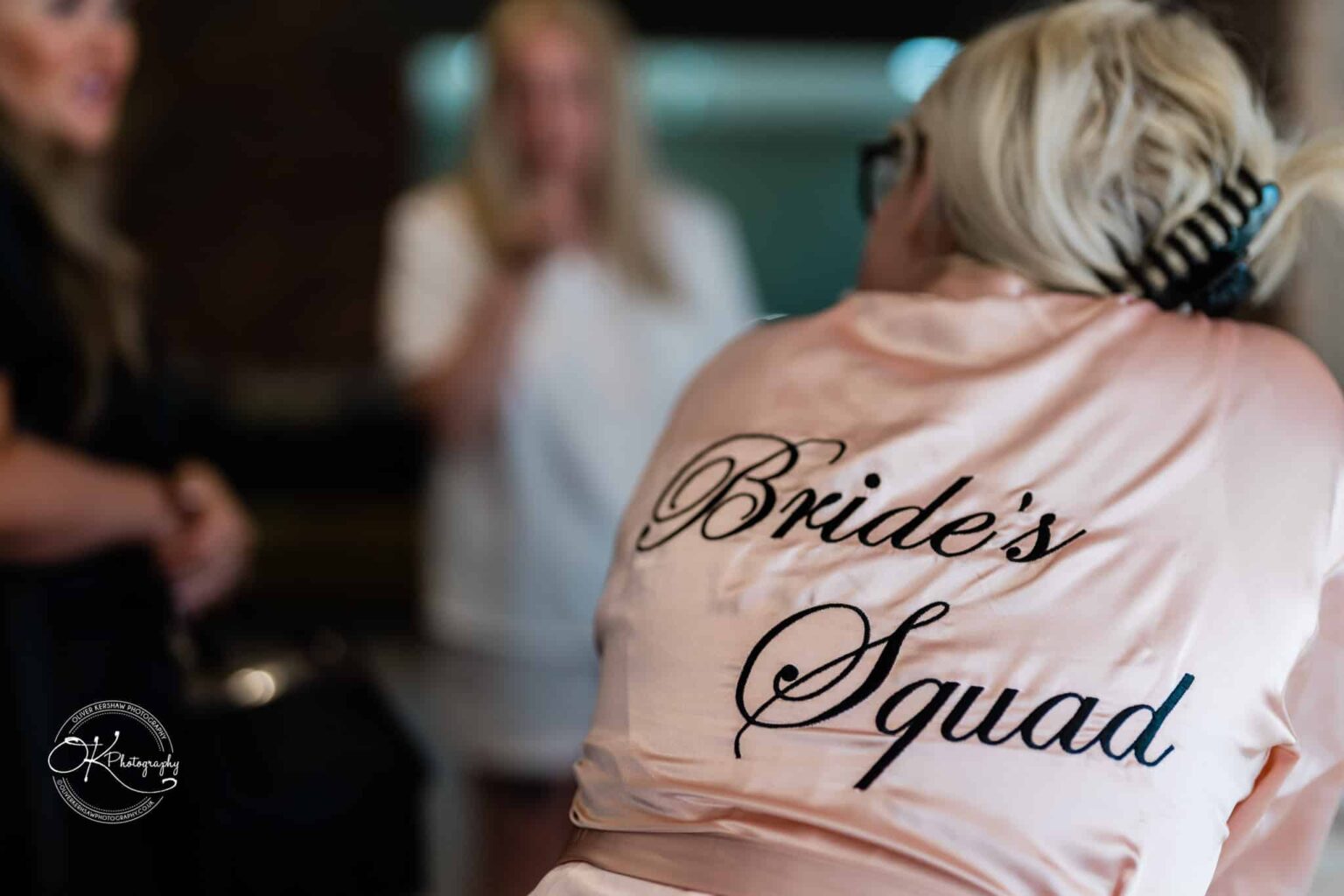 Person wearing a pink robe with "Bride's Squad" written on the back, with other people conversing in the background.
