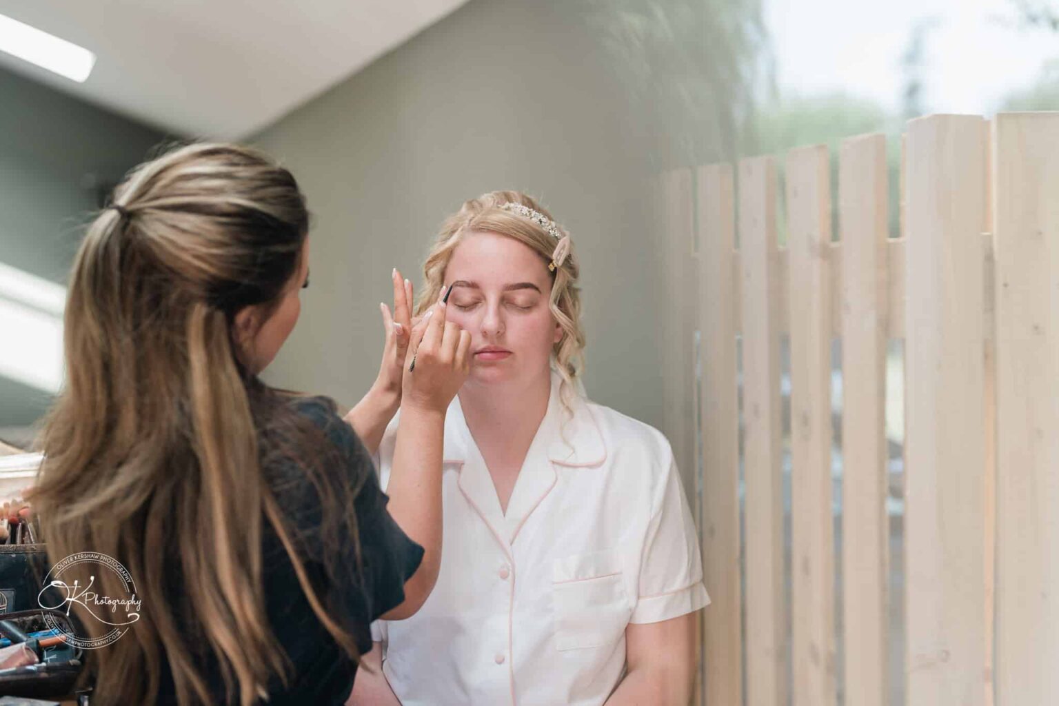 A woman having her makeup done, with another woman applying product to her eyebrows. The woman receiving makeup has her eyes closed and is wearing a white shirt with pink trim.