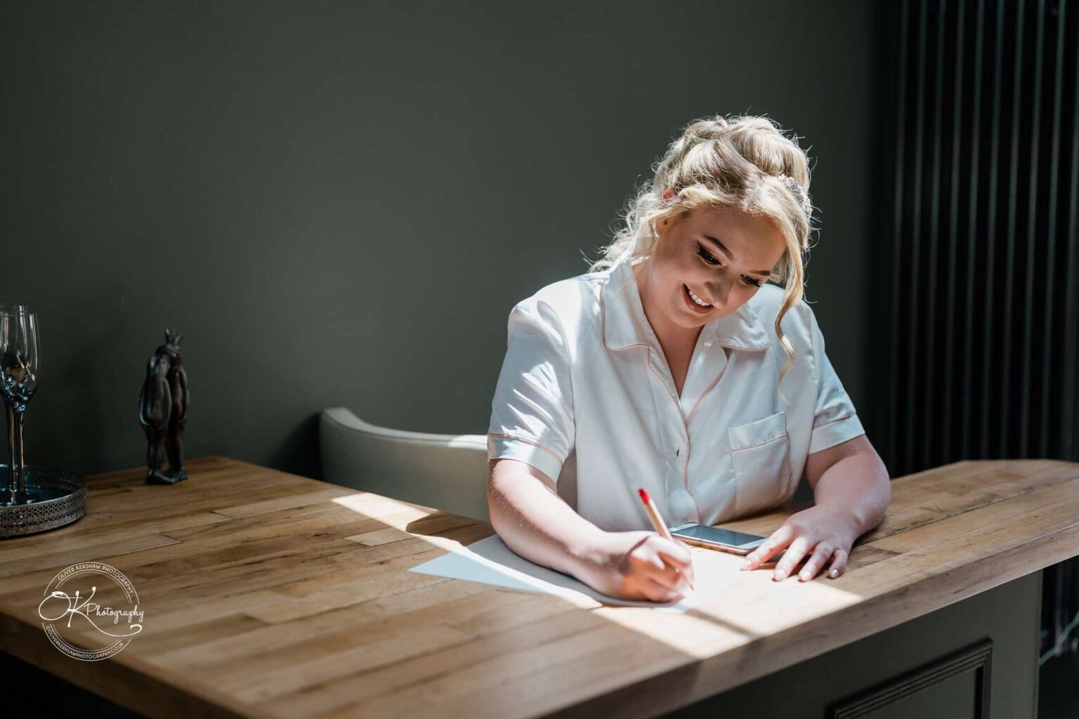 Woman smiling while writing at a wooden desk with a smartphone and empty glasses beside her.