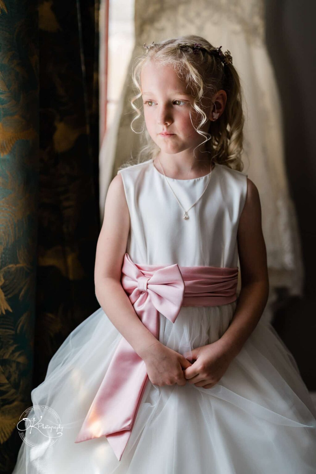 Young girl in a white dress with a pink sash and bow, looking towards the window.