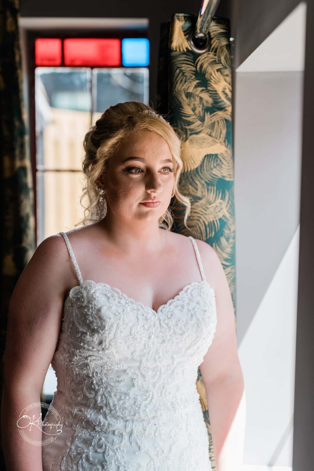 A bride in a white lace wedding dress standing near a window with colourful stained glass, looking out thoughtfully.