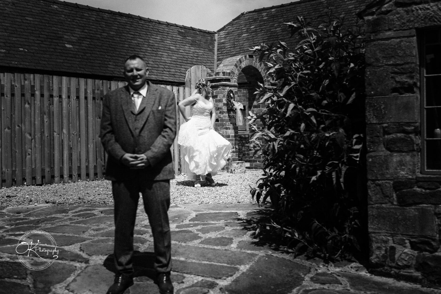 A groom standing in the foreground with his hands clasped, while a bride in a wedding dress walks behind him within a courtyard area, surrounded by rustic stone buildings and a wooden fence.