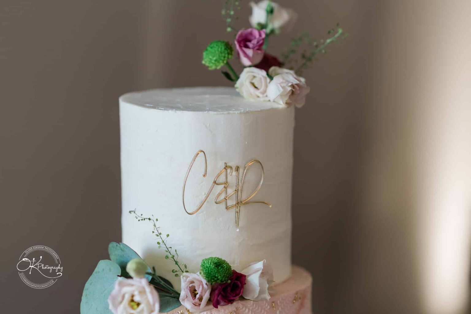 A white wedding cake with a gold wire monogram topper and floral decorations, featuring pink and white flowers and green accents.