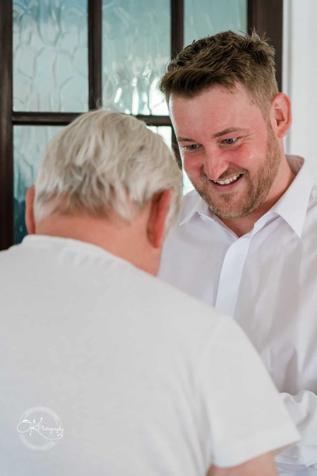 Two men, one with grey hair and one with short brown hair, sharing a moment together; the older man is facing away from the camera while the younger is smiling, dressed in a white shirt.