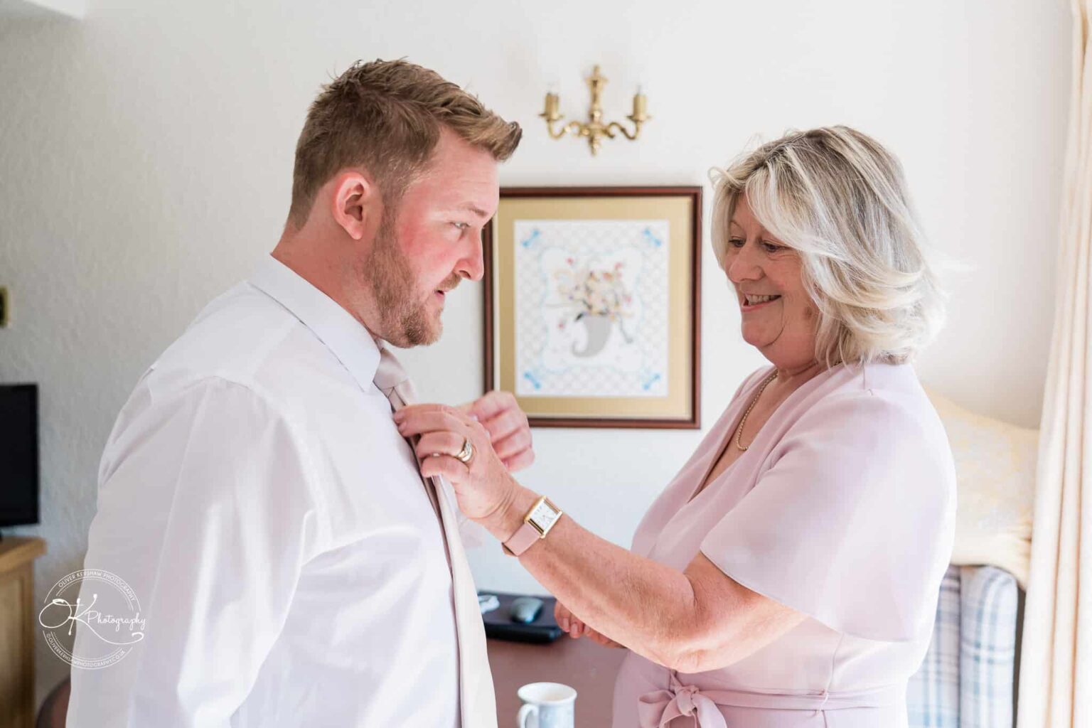 A woman fastening the tie of a man dressed in a white shirt, both smiling, in a warmly lit room.