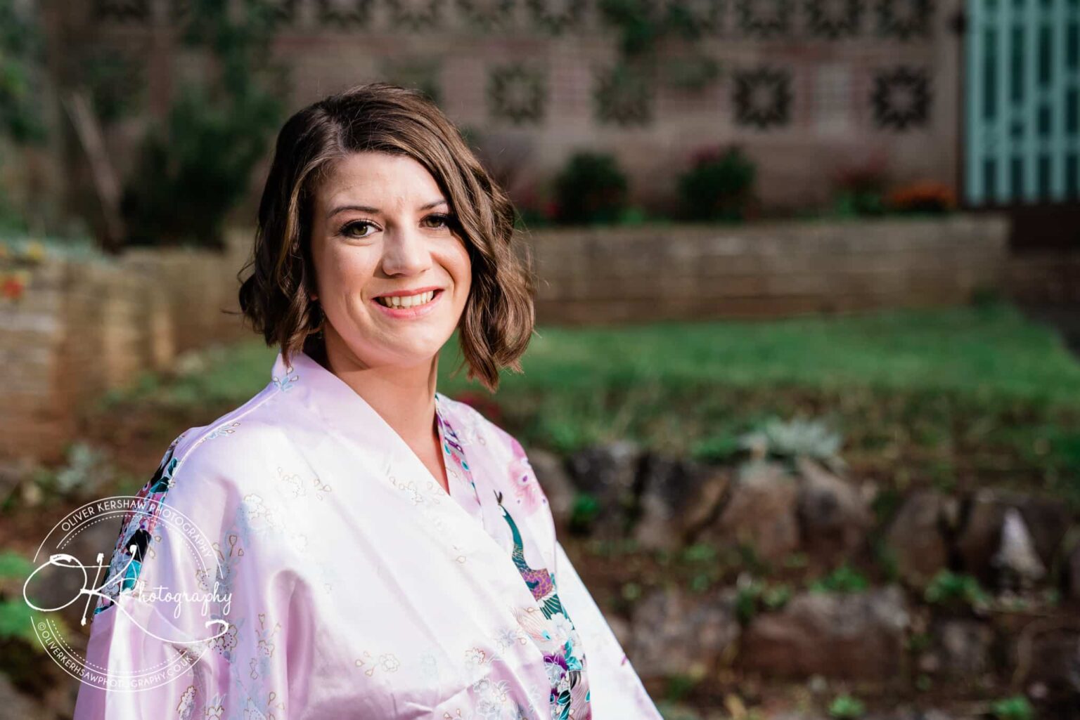 A smiling woman wearing a light purple robe, standing outdoors with a brick wall and greenery in the background.