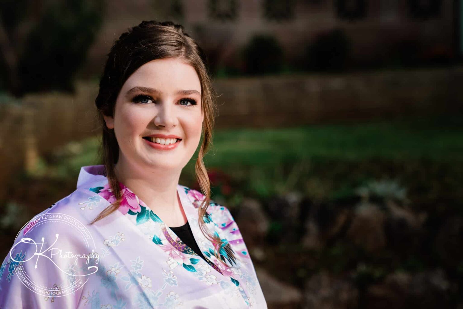 A woman in a floral robe smiling outdoors with soft greenery and a building in the background.