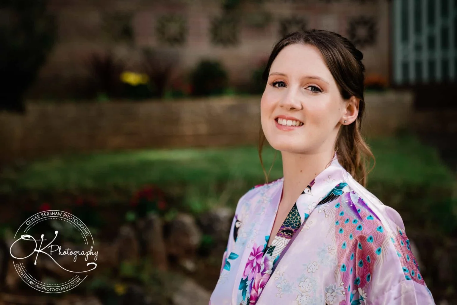 A woman in a purple floral robe smiling, with greenery and a brick wall in the background.
