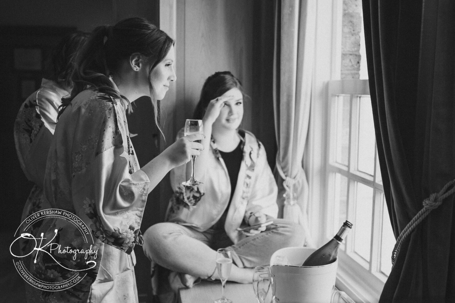 Two women in robes, one holding a glass of champagne, looking out a window with a champagne bottle in an ice bucket nearby.