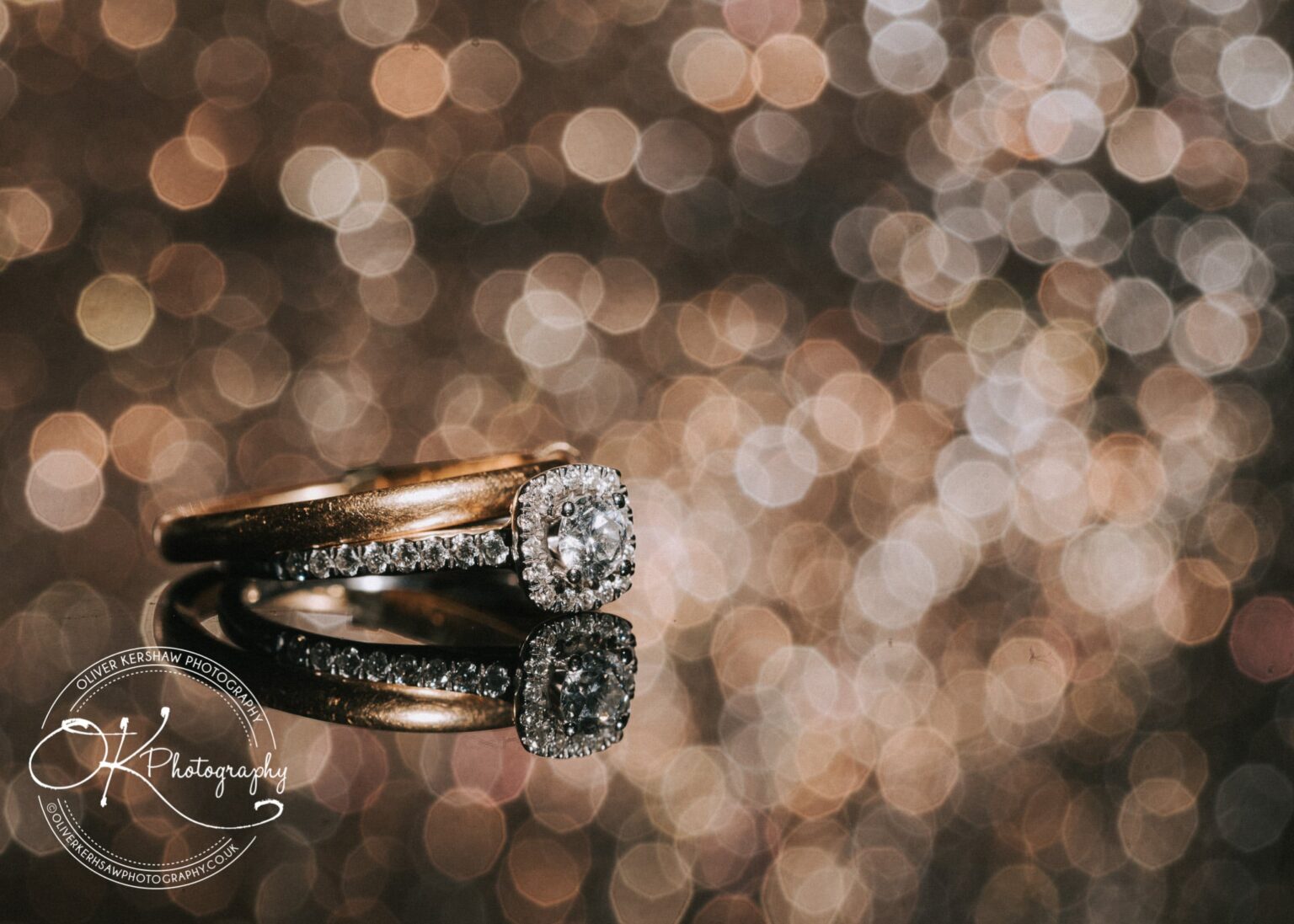 Close-up of a diamond engagement ring and wedding band on a reflective surface with a sparkling bokeh background.