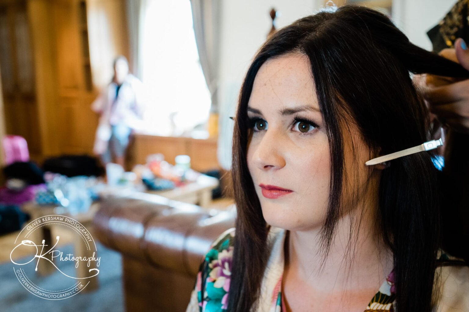 A woman getting her hair styled at a salon with a blurred background showing a person standing near a window.