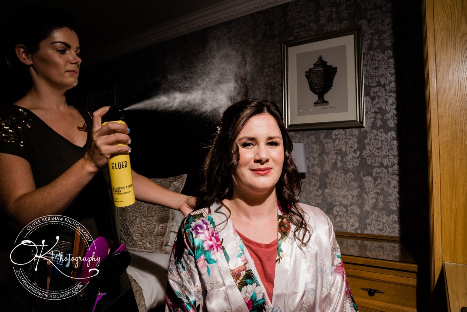 A woman in a floral robe having her hair styled with hairspray by a makeup artist in a dimly lit room.