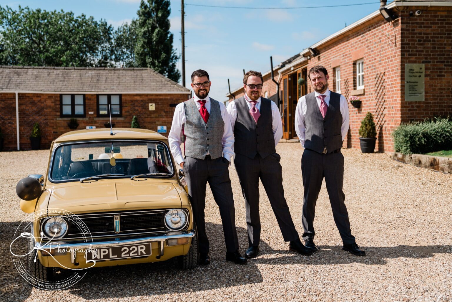 Three men in suits and waistcoats with red ties standing beside a vintage yellow car, posing for a photograph outdoors in front of brick buildings.