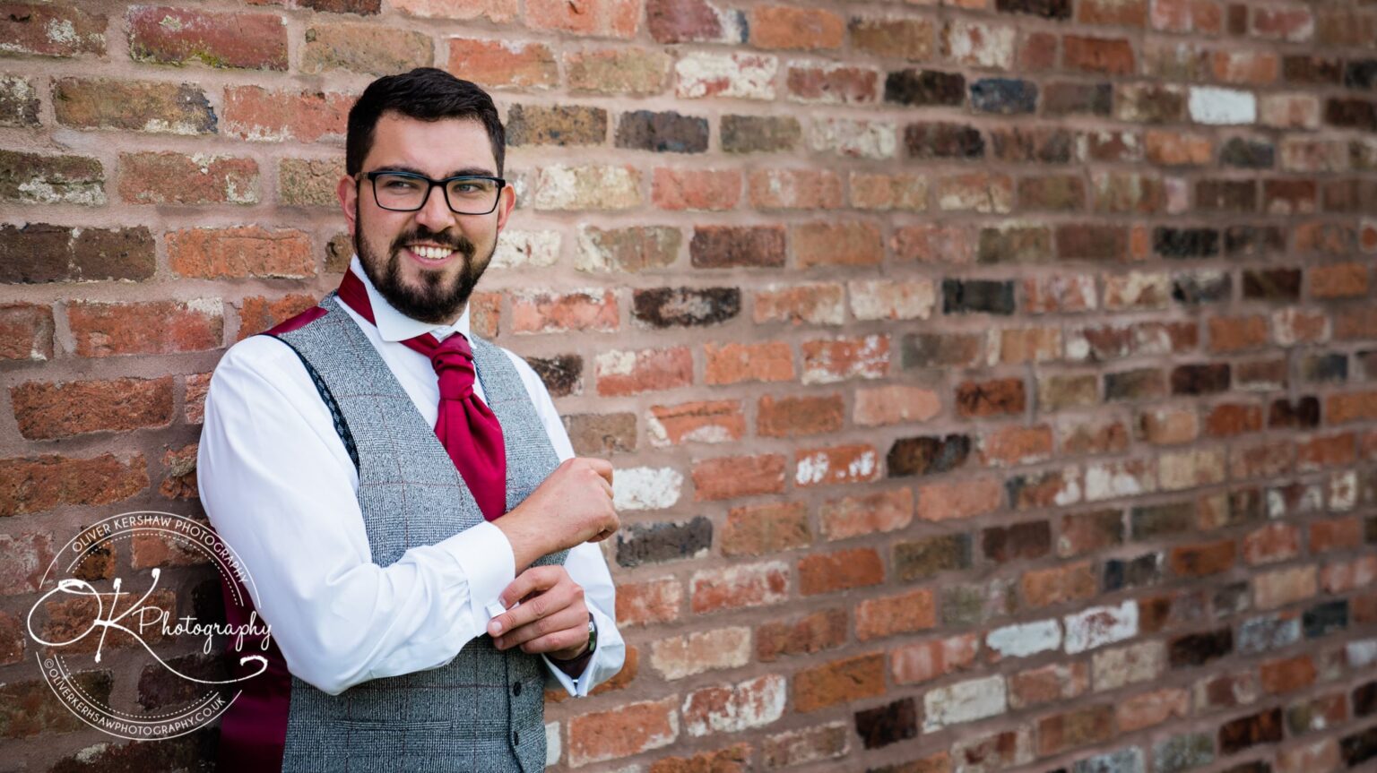 Groom posing against a brick wall, wearing a grey waistcoat, white shirt, red tie, and glasses with a smile on his face.