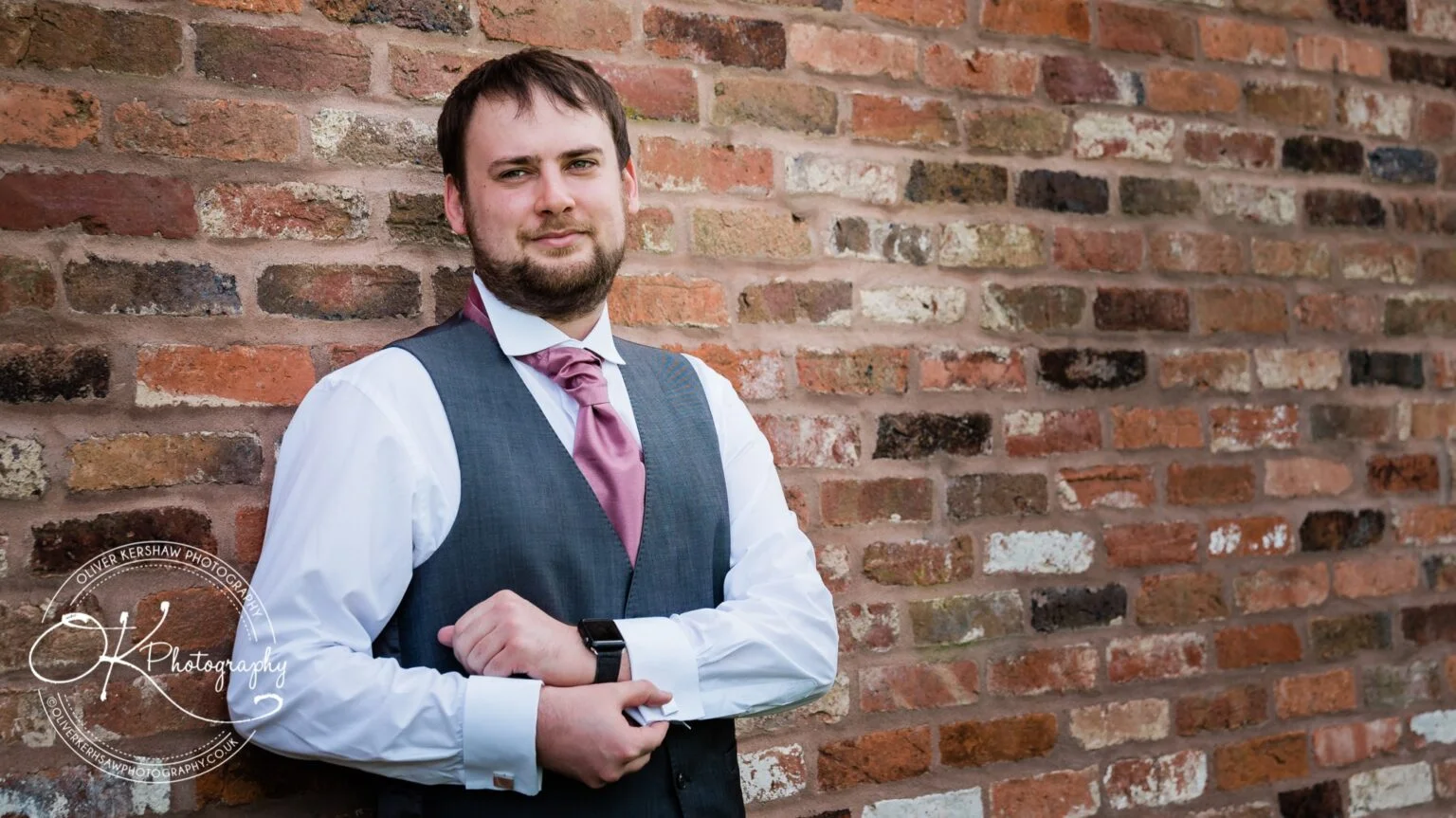 A man in a waistcoat and tie leans against a rustic brick wall, posing for a wedding photograph.