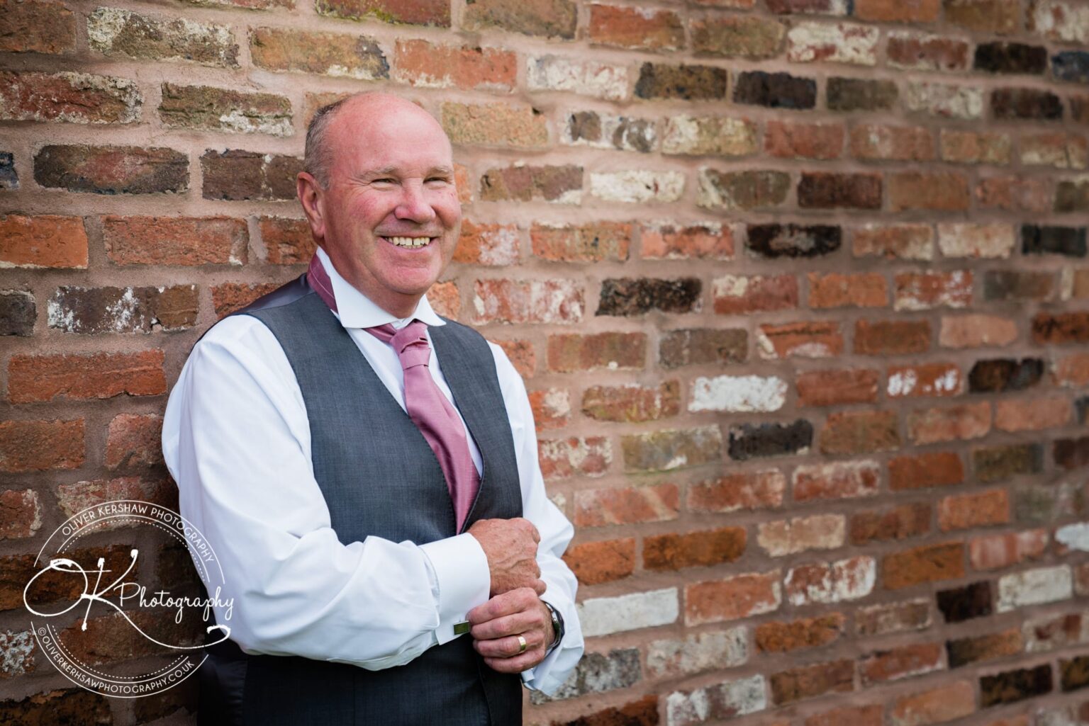 A smiling man in a suit and pink tie leaning against a brick wall.