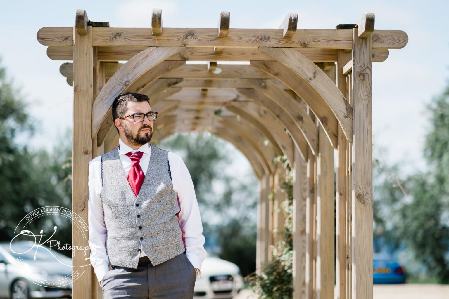 Man in formal attire standing under a wooden pergola at a wedding venue.