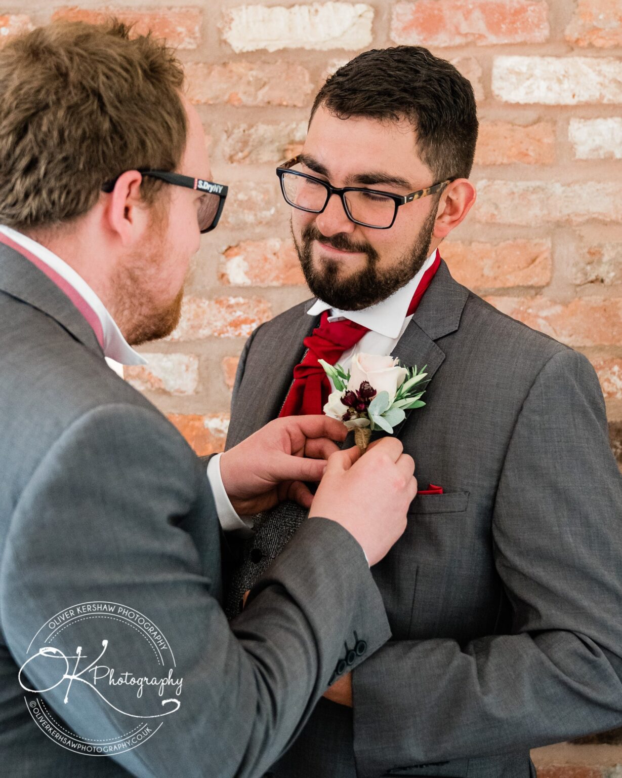 A groom being assisted with his boutonniere by another man against a brick wall backdrop.