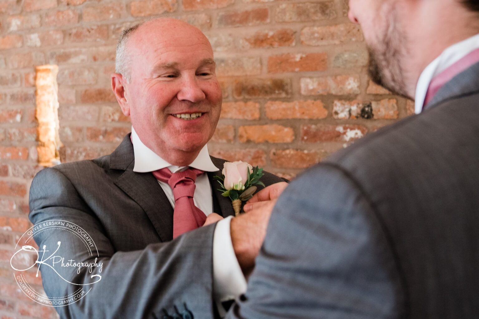 A man smiling while another man adjusts his boutonnière against a brick wall background.
