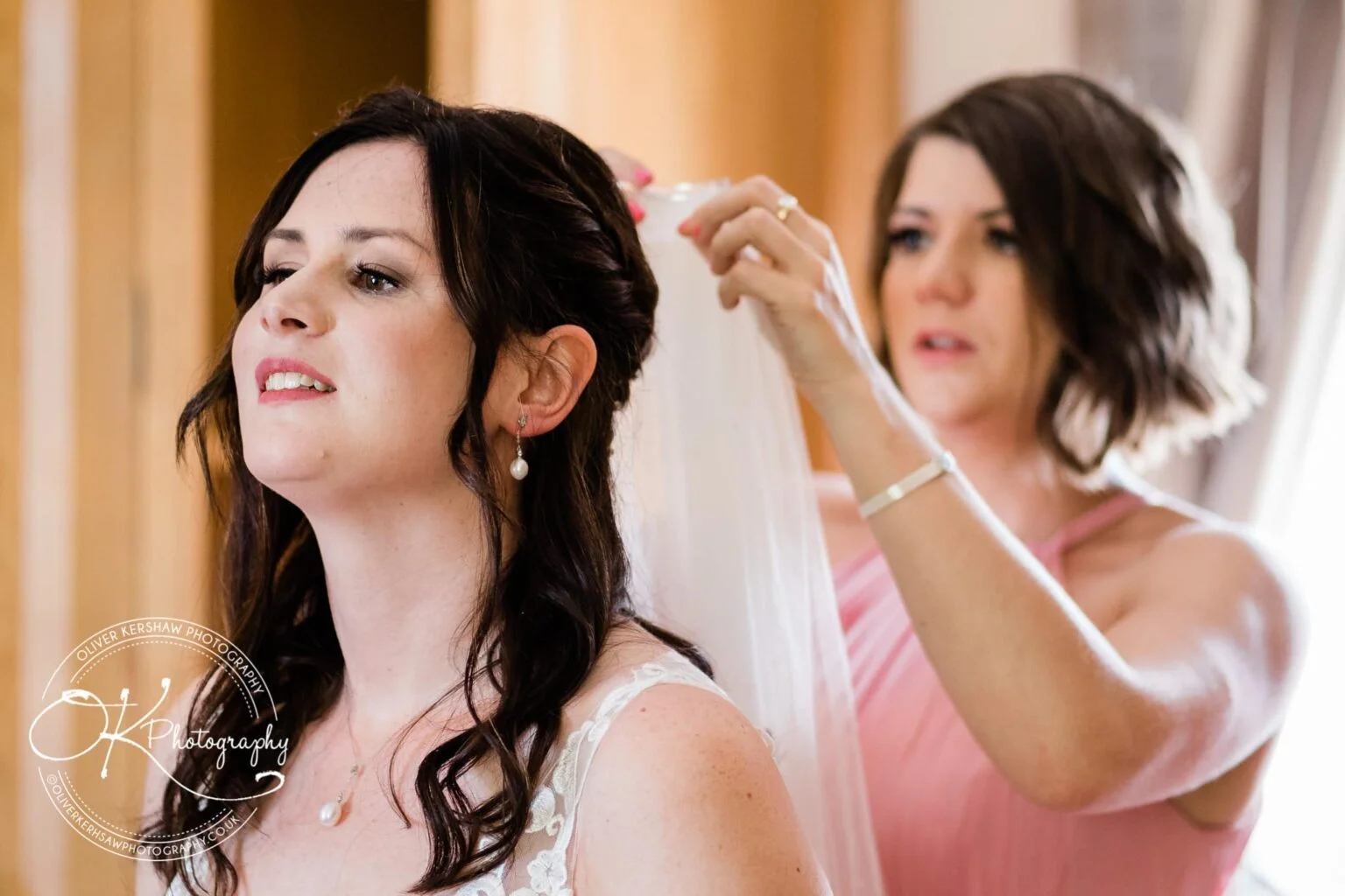 A woman helps another woman, dressed in wedding attire, adjust her veil.