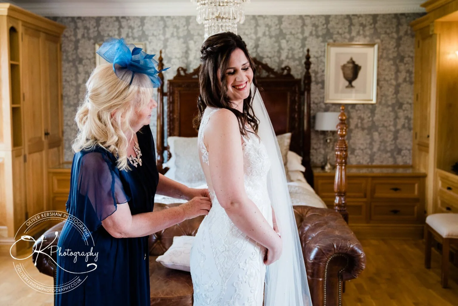 Bride smiles while having her wedding dress adjusted by a woman in a blue dress and hat in a luxurious bedroom.