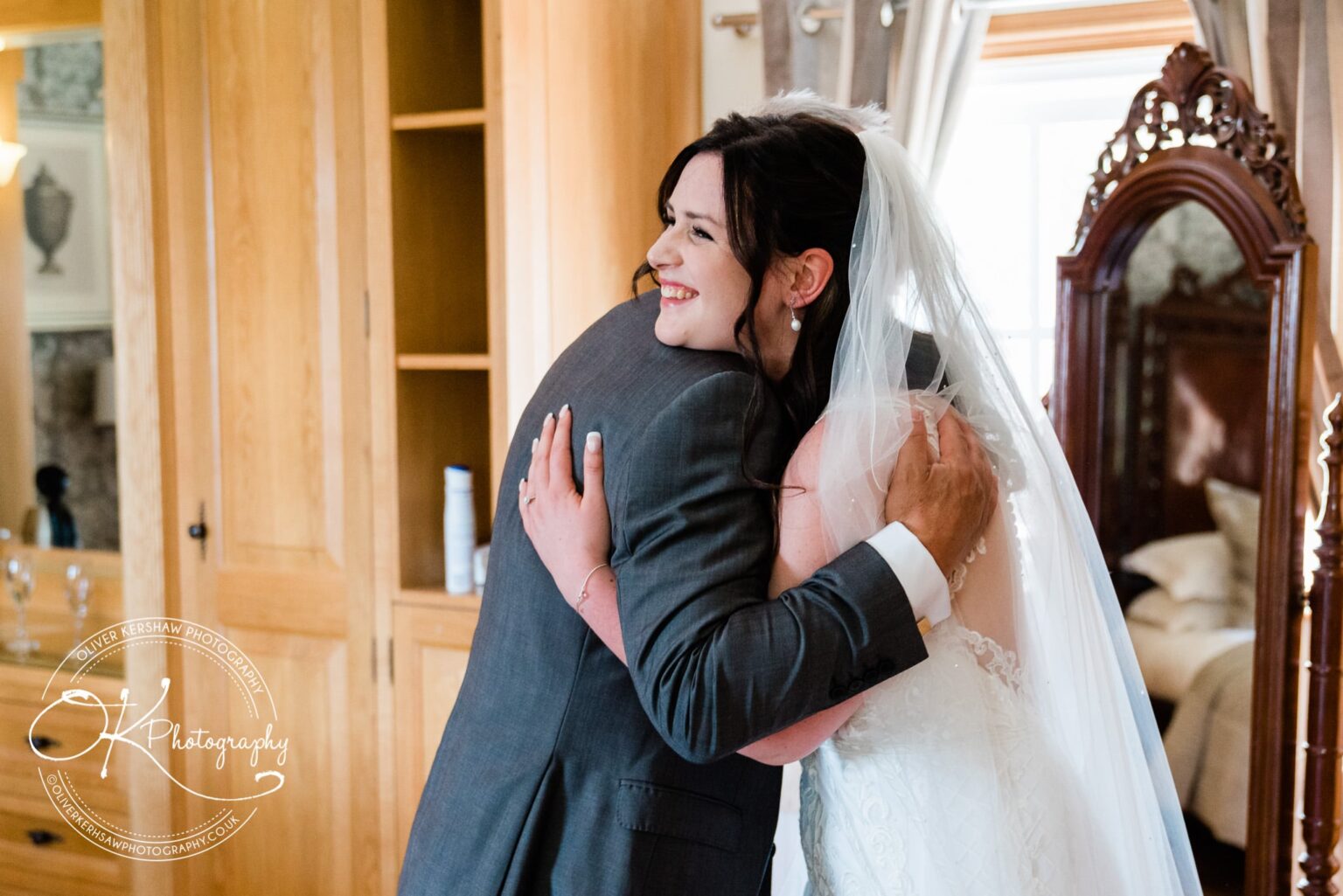 A bride in a white wedding dress and veil, smiling and hugging a man in a suit in an indoor setting.