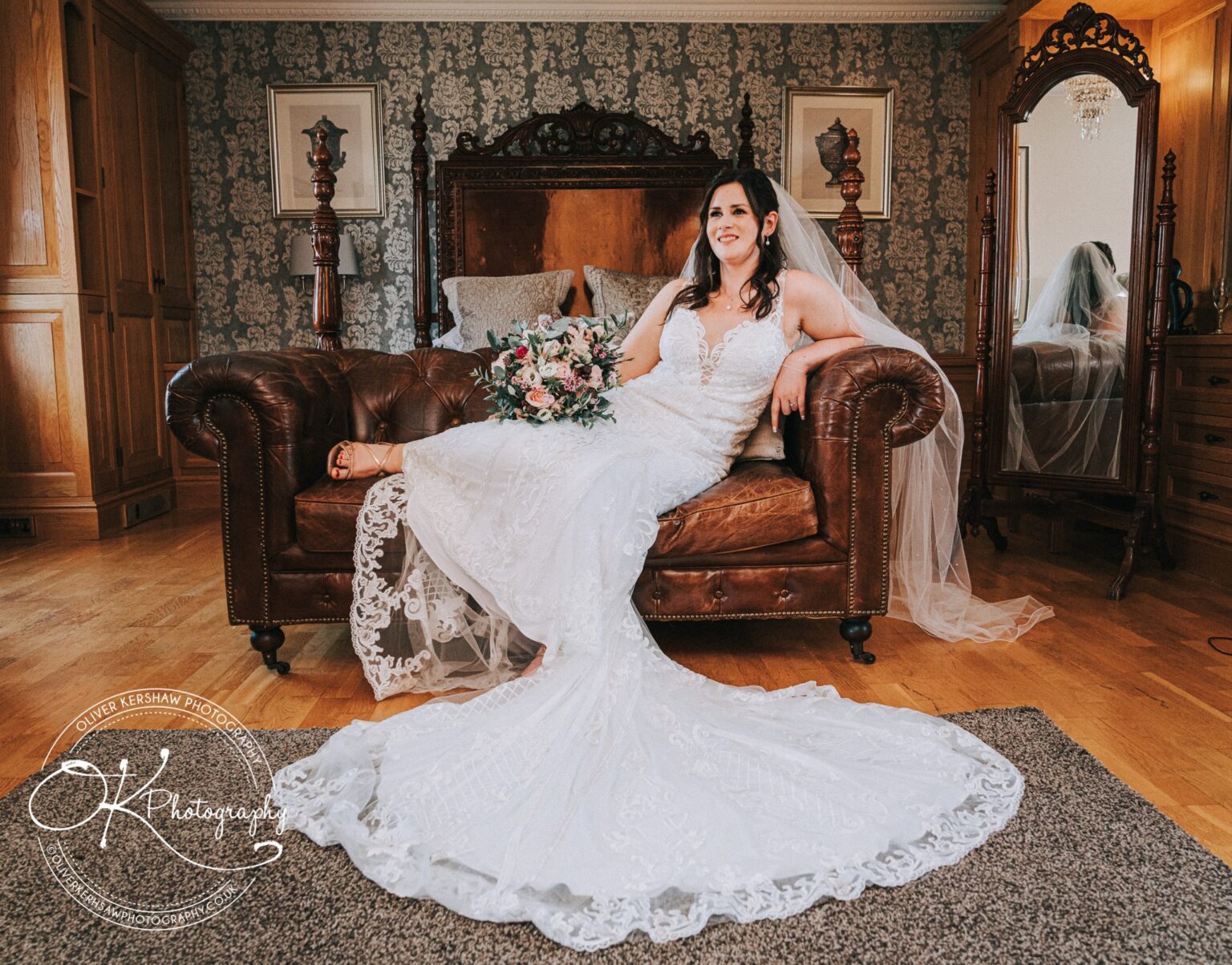Bride in a white lace wedding dress lounging on a brown leather sofa, holding a bouquet with a long veil trailing on the floor in a decorated room.