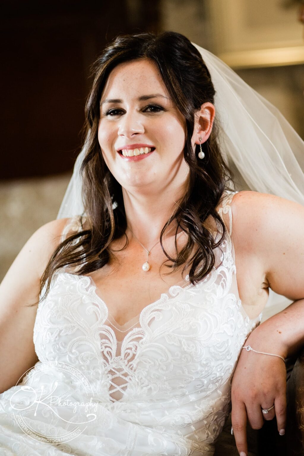 Bride in a white lace wedding gown and veil, smiling while seated indoors.