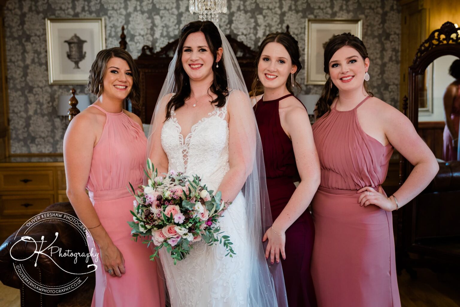 Bride in a white wedding dress surrounded by bridesmaids in pink and burgundy dresses, holding a bouquet, with a decorative indoor background.