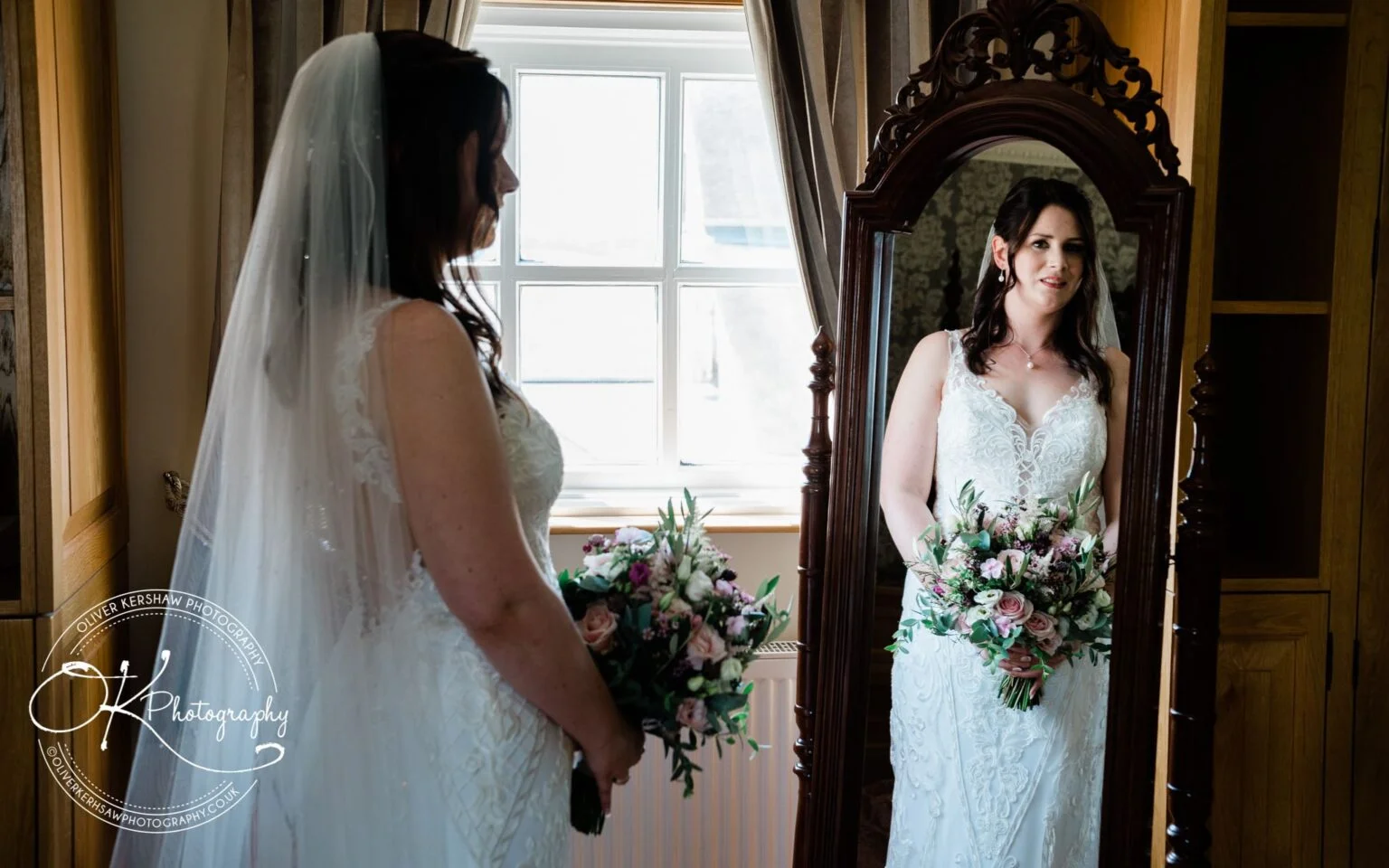 Bride in a white wedding dress holding a bouquet, looking at her reflection in a full-length mirror.