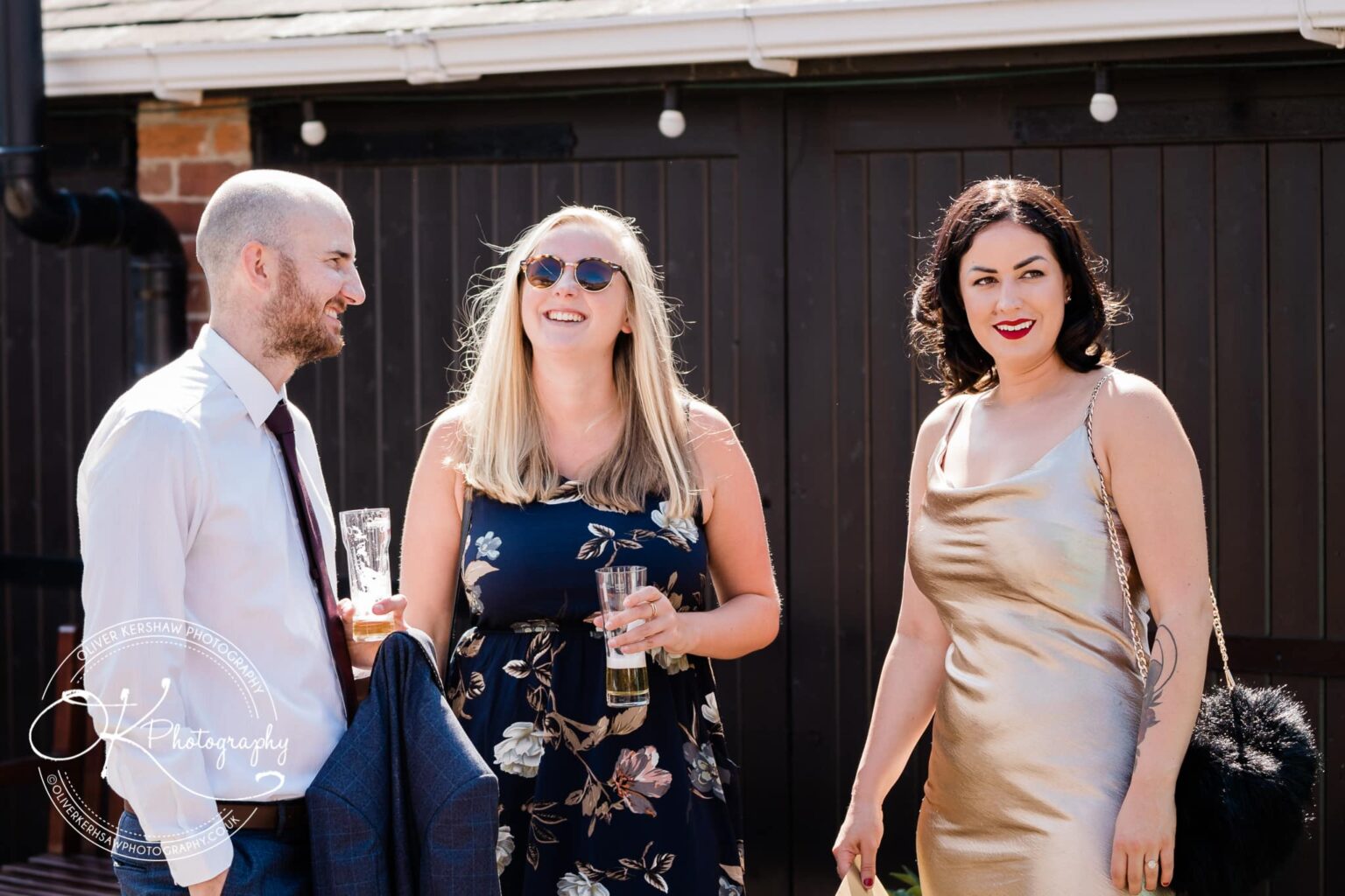 Three people at a wedding, two women and one man, smiling and holding drinks, outdoors.