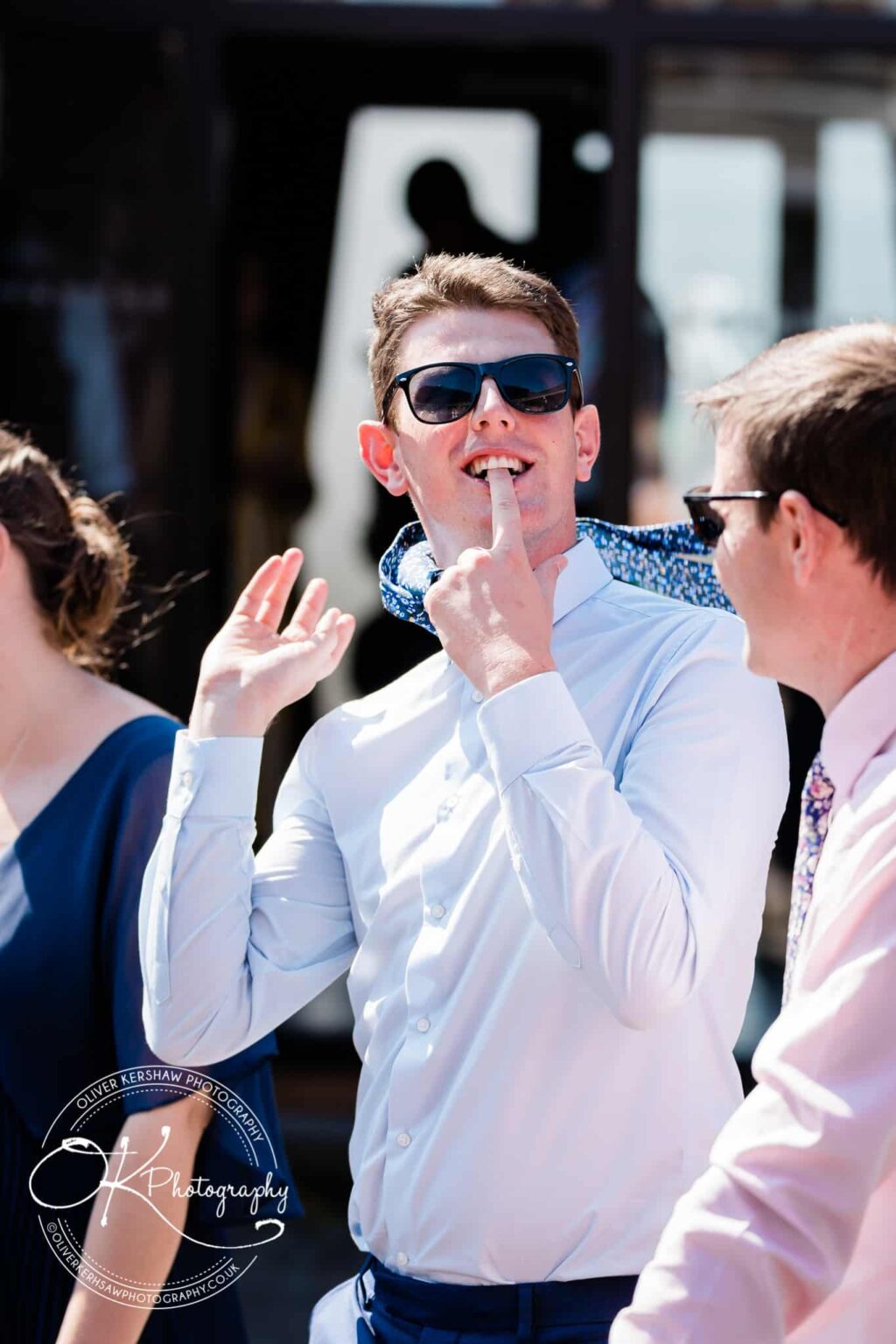 A man wearing sunglasses and a white shirt poses playfully while standing next to other people outdoors.