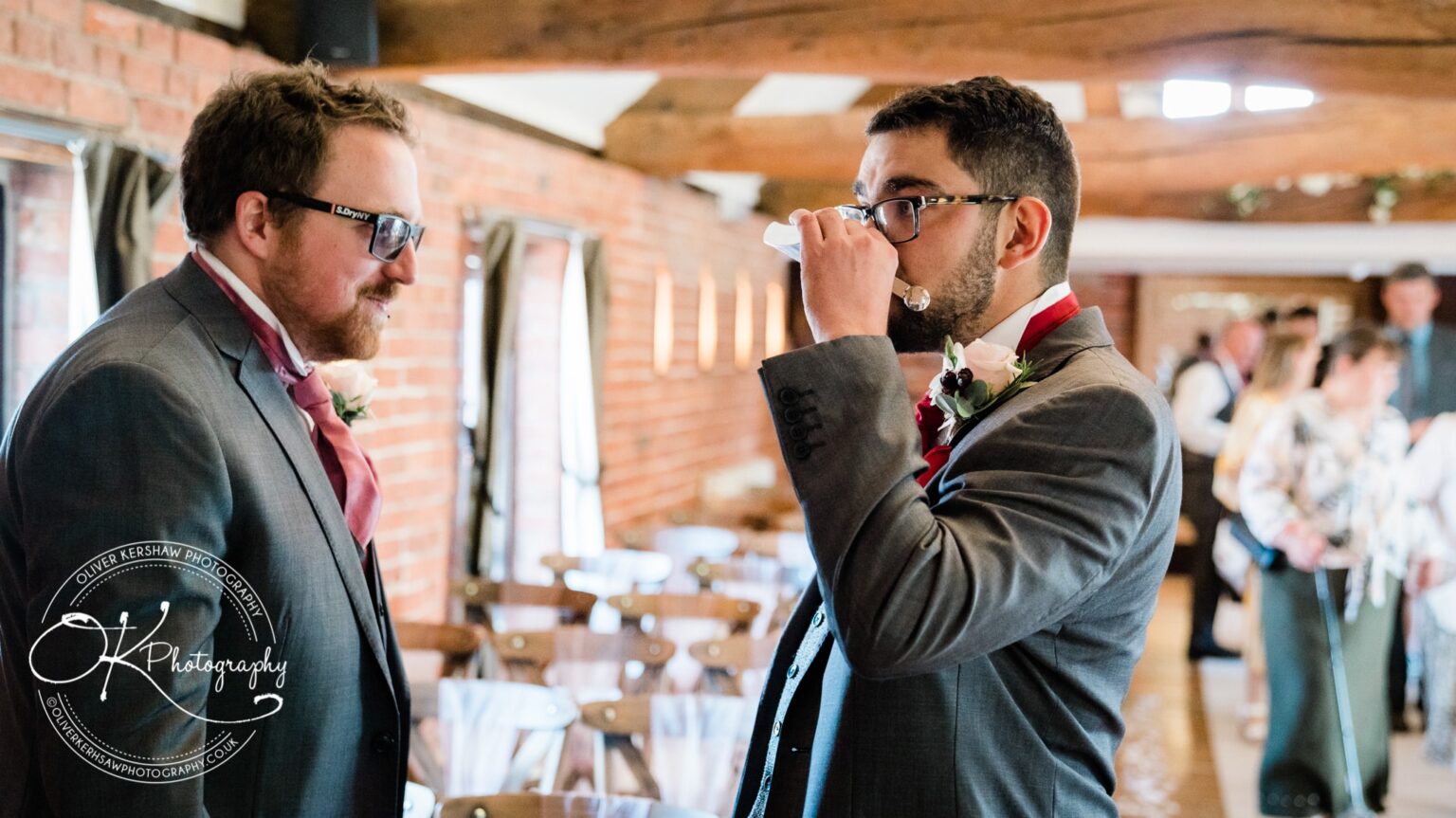 Two men in suits with boutonnieres at a wedding reception, one drinking from a glass, at a venue with brick walls and wooden beams.