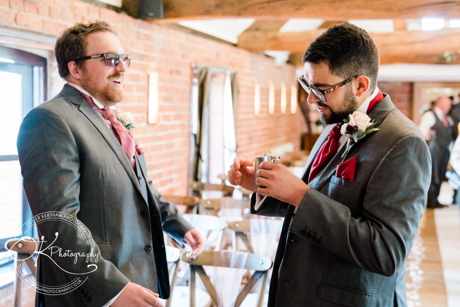 Two men in suits and red cravats, one holding a hip flask, laughing and talking in a rustic wedding venue with exposed brick walls.