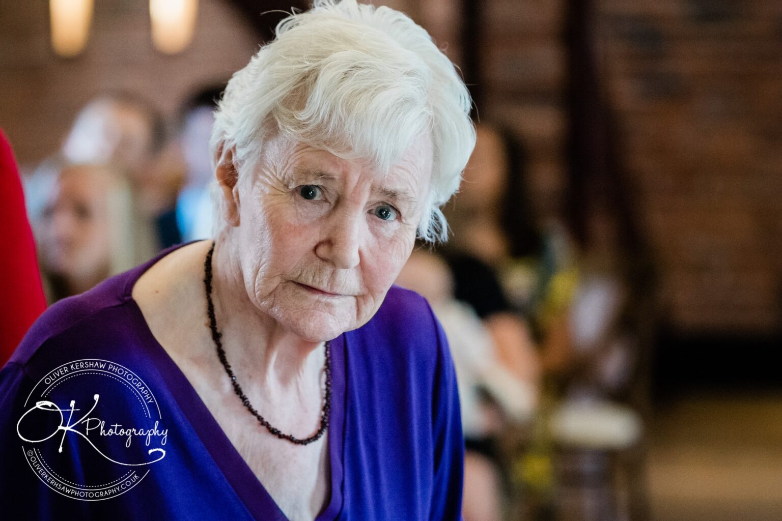 An elderly woman with short white hair, wearing a purple top and a beaded necklace, looking at the camera with a serious expression; background shows blurred figures and a softly lit room.