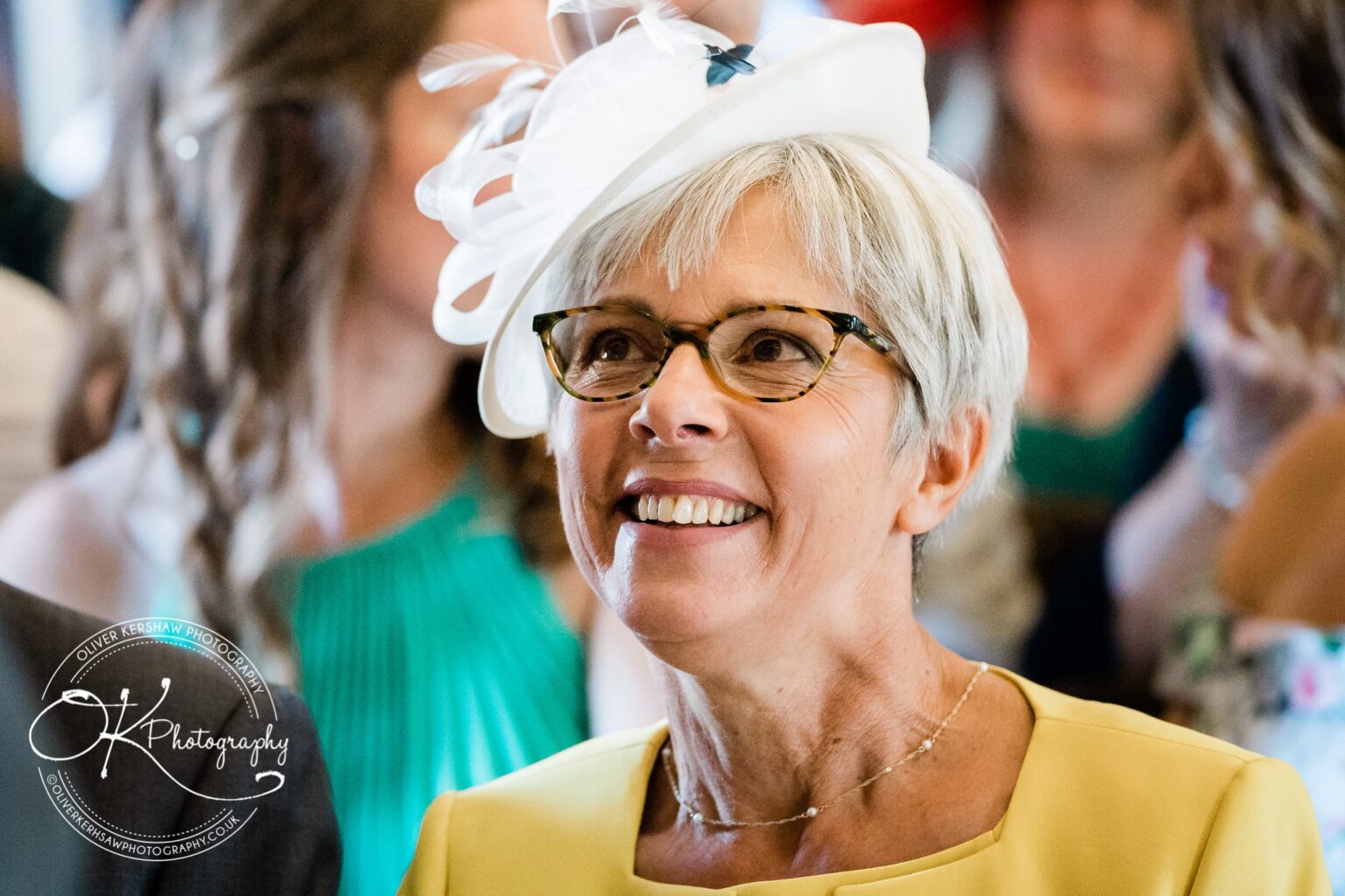 An older woman wearing glasses and a white fascinator, smiling in a crowd at a wedding.