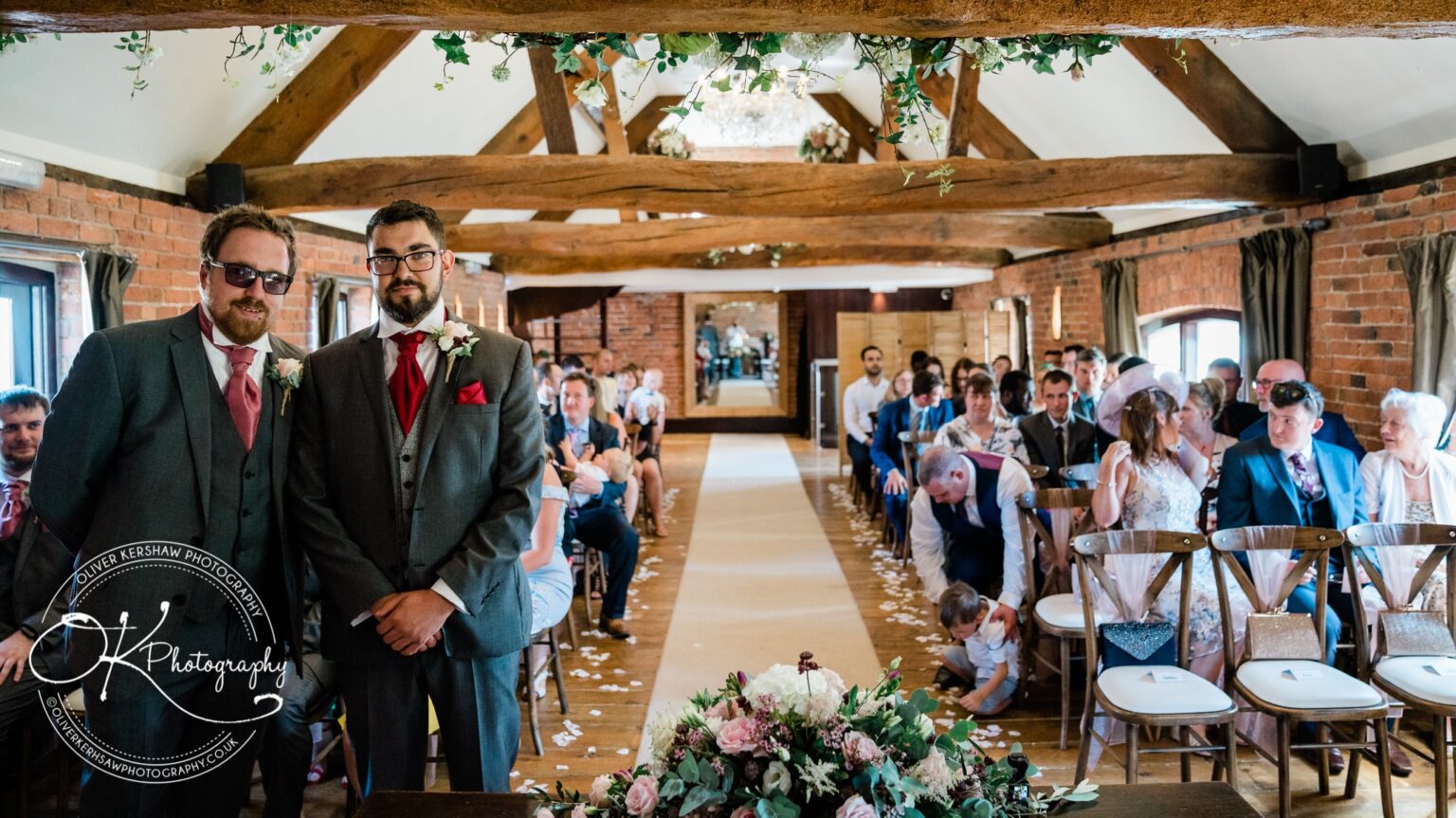 Two groomsmen standing at the altar in a rustic barn wedding venue, with guests seated on both sides.