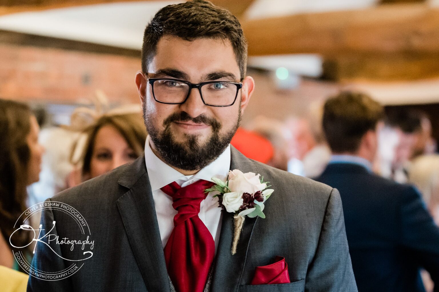 Man in a suit with a red tie and boutonnière, smiling at the camera with a blurred crowd in the background.