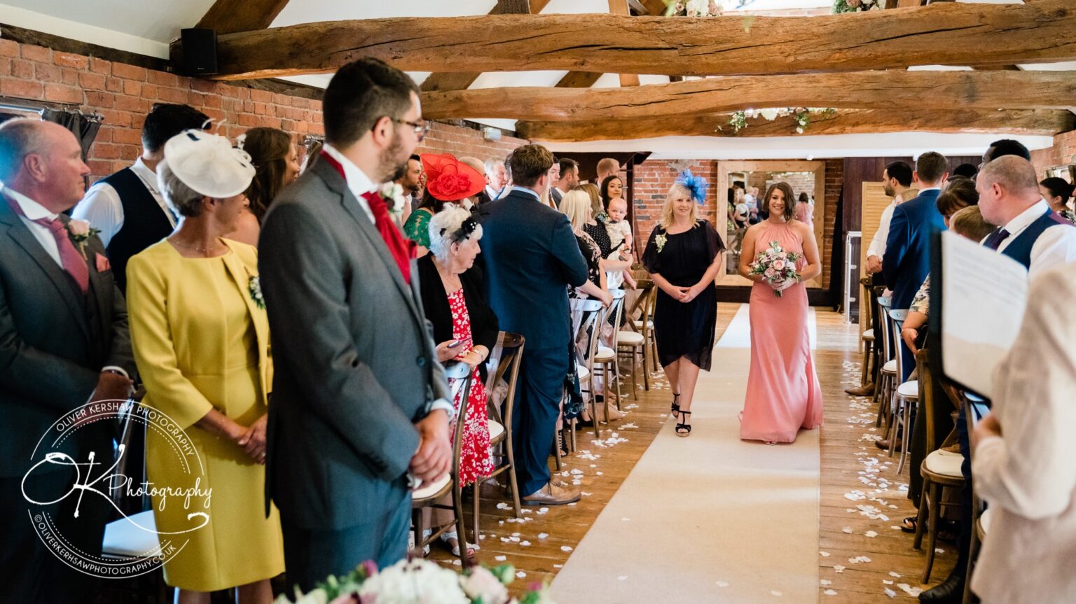 Guests seated in a rustic venue watching a wedding procession with bridesmaids walking down the aisle.
