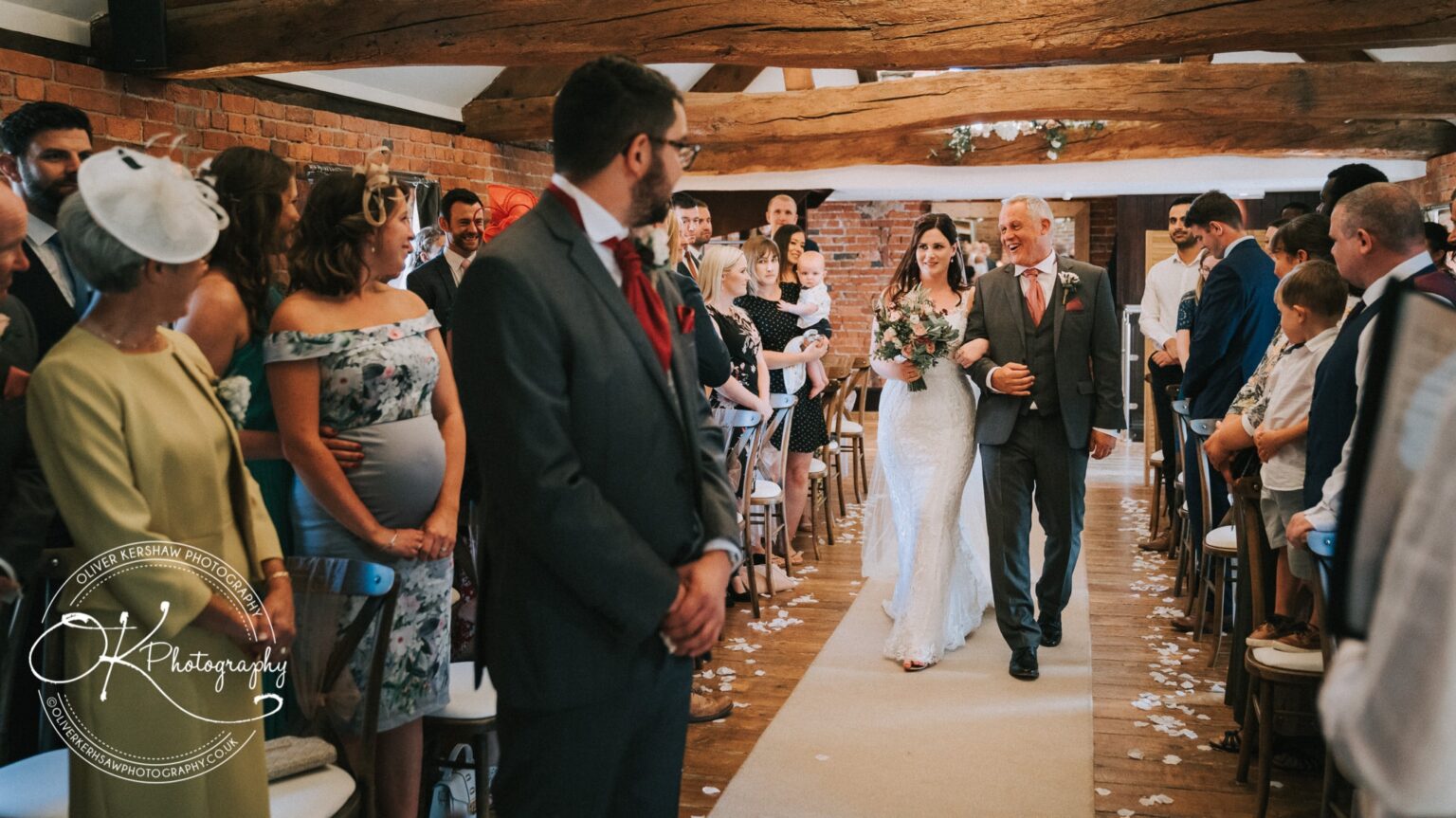 Bride walking down the aisle with her father, surrounded by guests at an indoor wedding ceremony with exposed wood beams and brick walls.