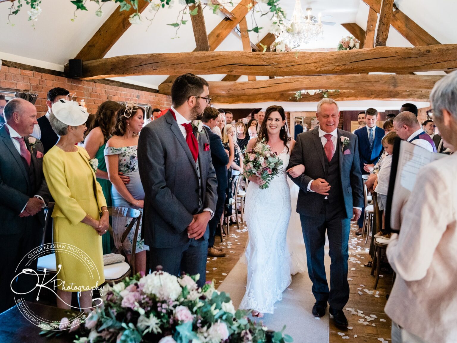 A bride walks down the aisle with an older man at Swancar Farm, surrounded by seated guests in a rustic setting with exposed wooden beams and floral decorations.