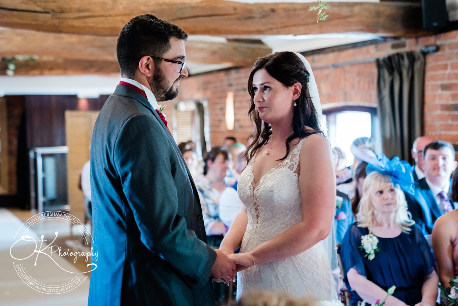 Bride and groom holding hands and looking at each other during a wedding ceremony at Swancar Farm, with guests seated in the background.