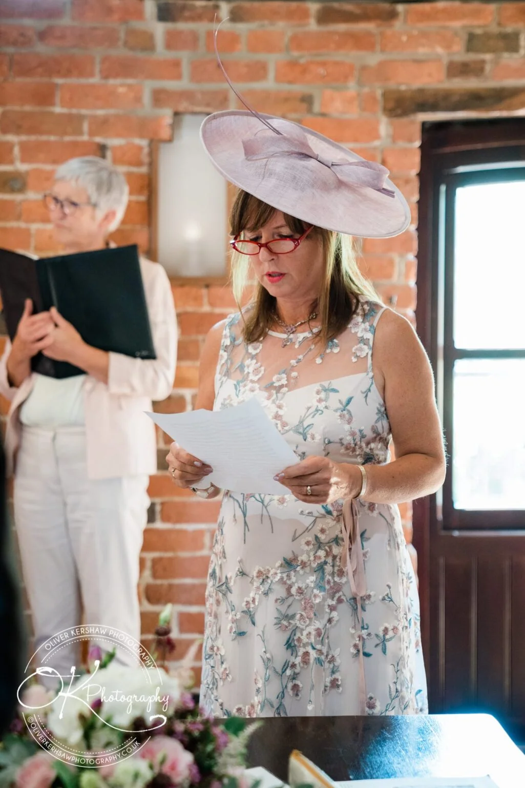 Woman in a detailed floral dress and large lavender hat reading a paper; another person in the background holding a book, with exposed brick walls behind them.