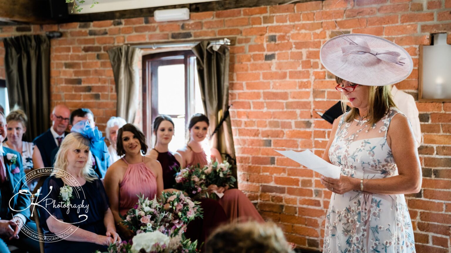 A woman in a floral dress and hat reads from a paper, while seated guests, including women holding bouquets, look on attentively in a brick-walled room.
