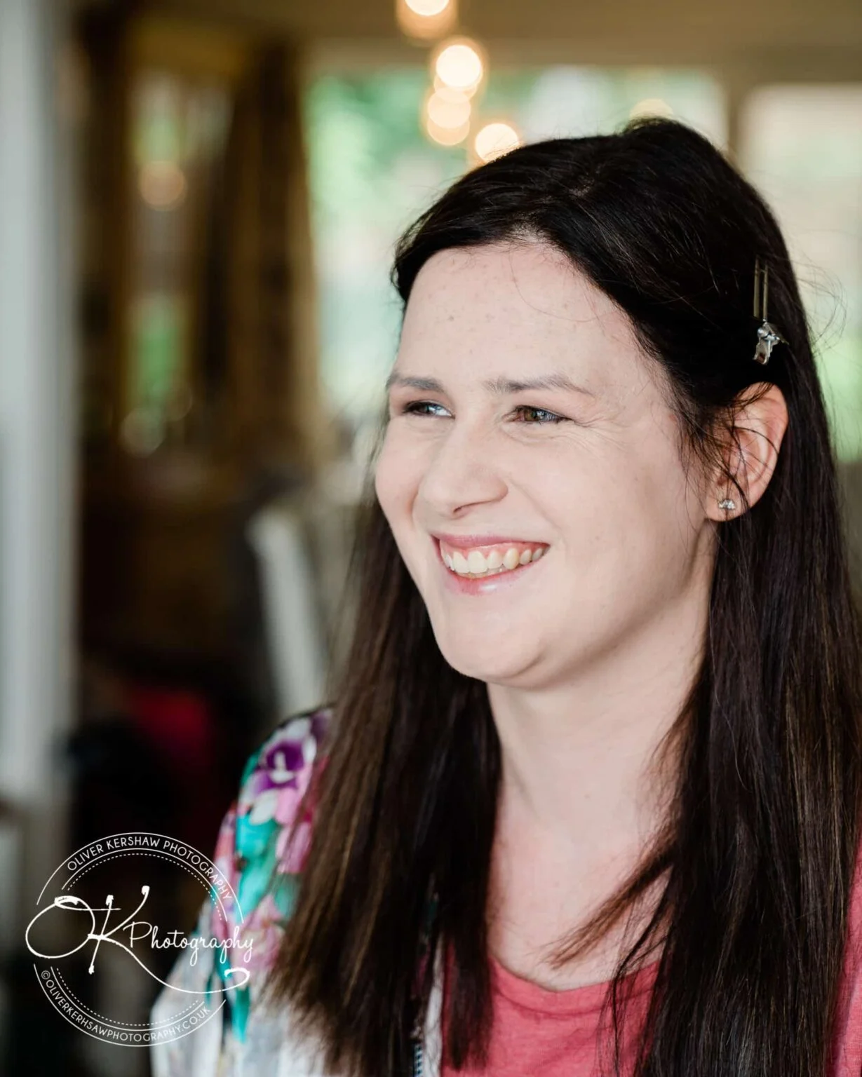A smiling person with long dark hair, wearing a pink top and floral jacket, with a blurred background and visible bokeh lights.