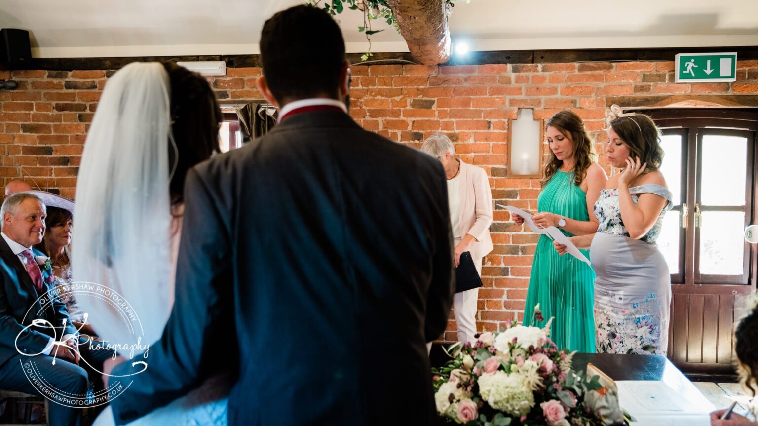 A wedding ceremony with two women reading from papers, a bride with a veil, a groom, and seated guests against a brick wall background.