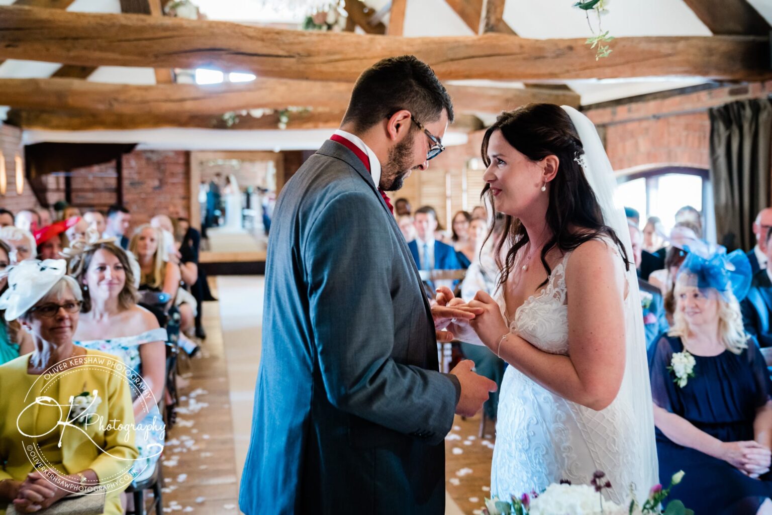 Bride and groom exchanging rings during their wedding ceremony at Swancar Farm, with guests watching in the background.