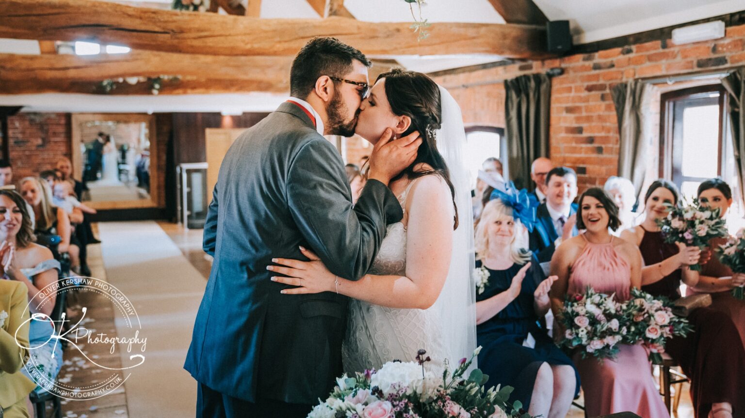 Bride and groom sharing a kiss during their wedding ceremony, surrounded by cheering family and friends in a rustic venue.