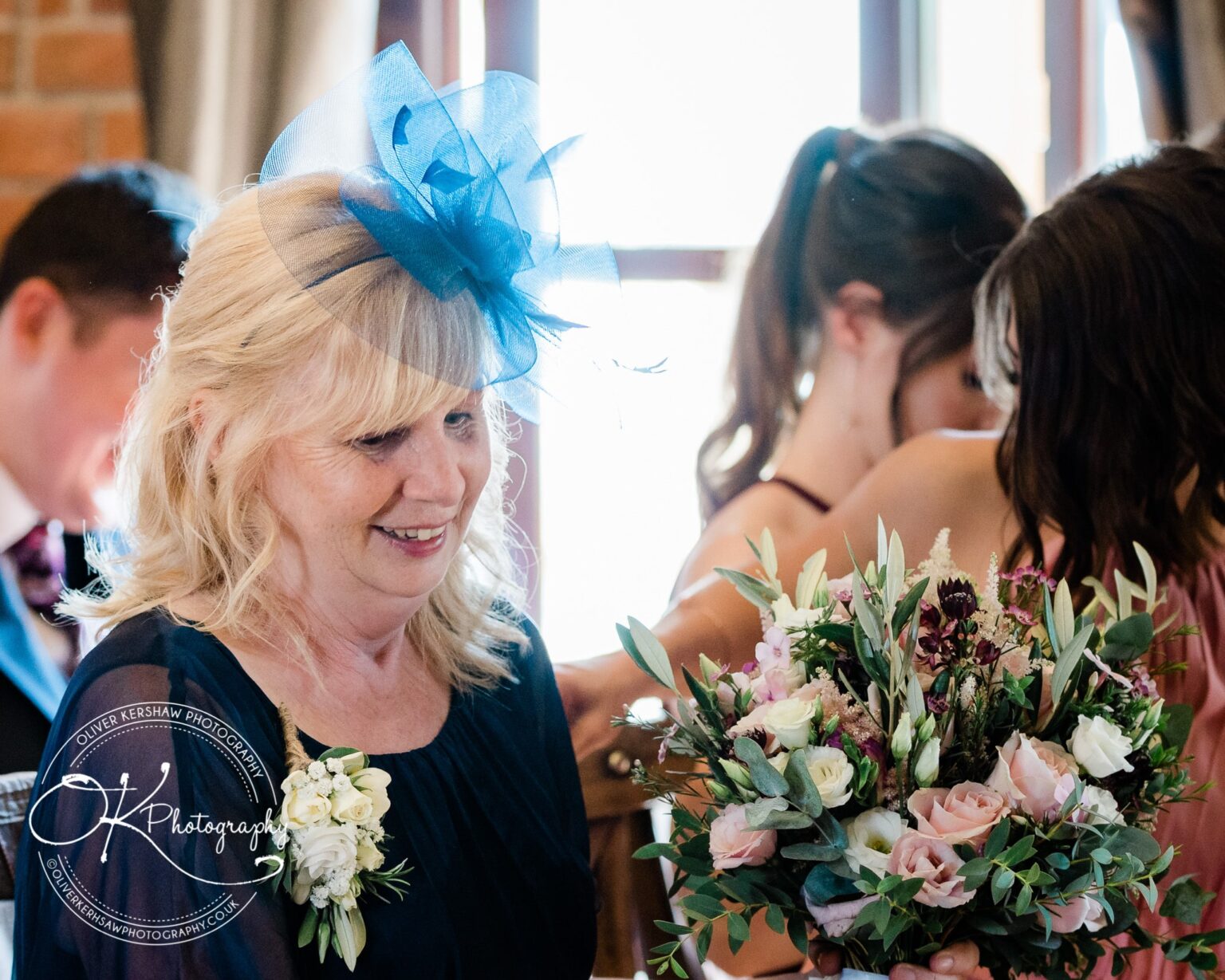 Blonde woman wearing a blue fascinator and holding a floral bouquet at an indoor event.